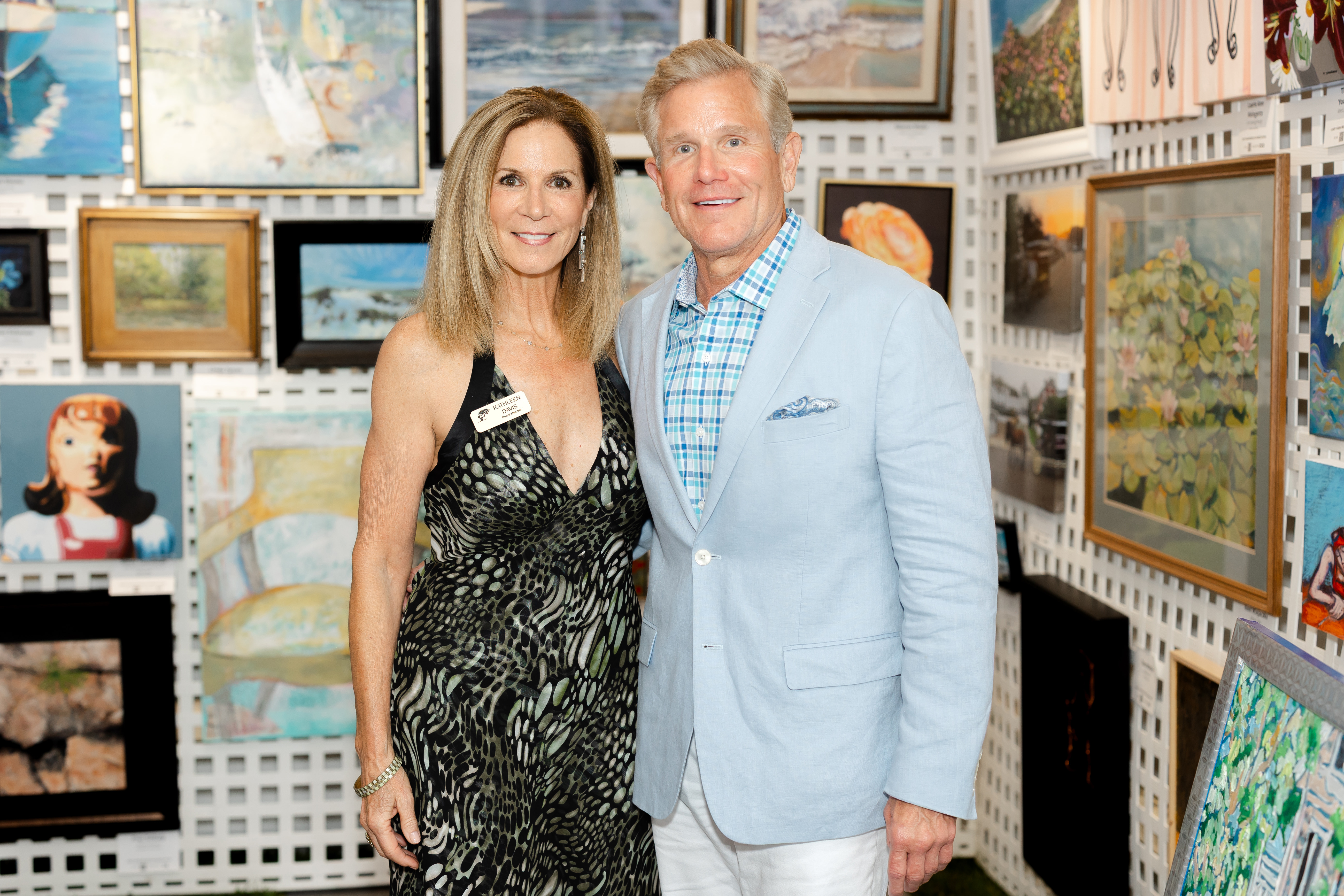 A smiling woman in a black dress standing next to her husband posing in front of an art gallery wall
