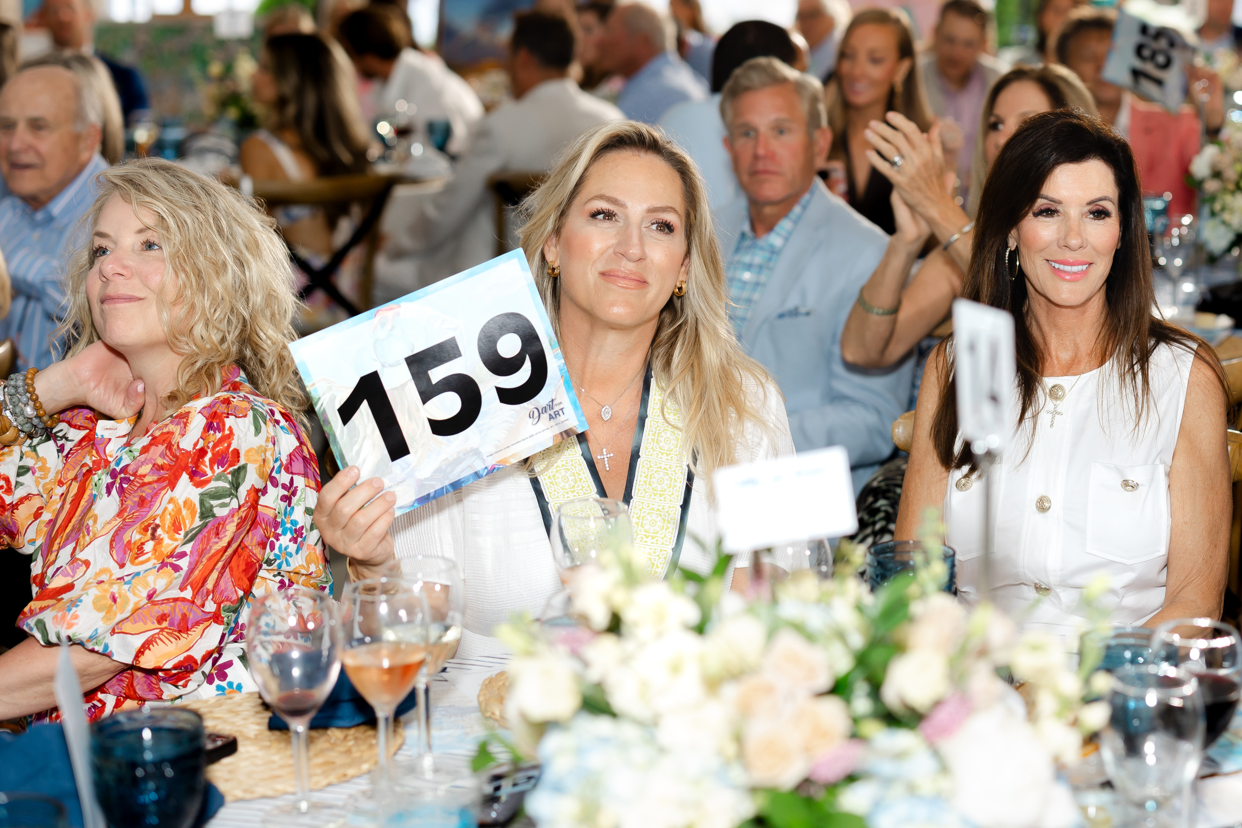 Three elegant women seated at a decorated event dining table holding up their bidder paddles.