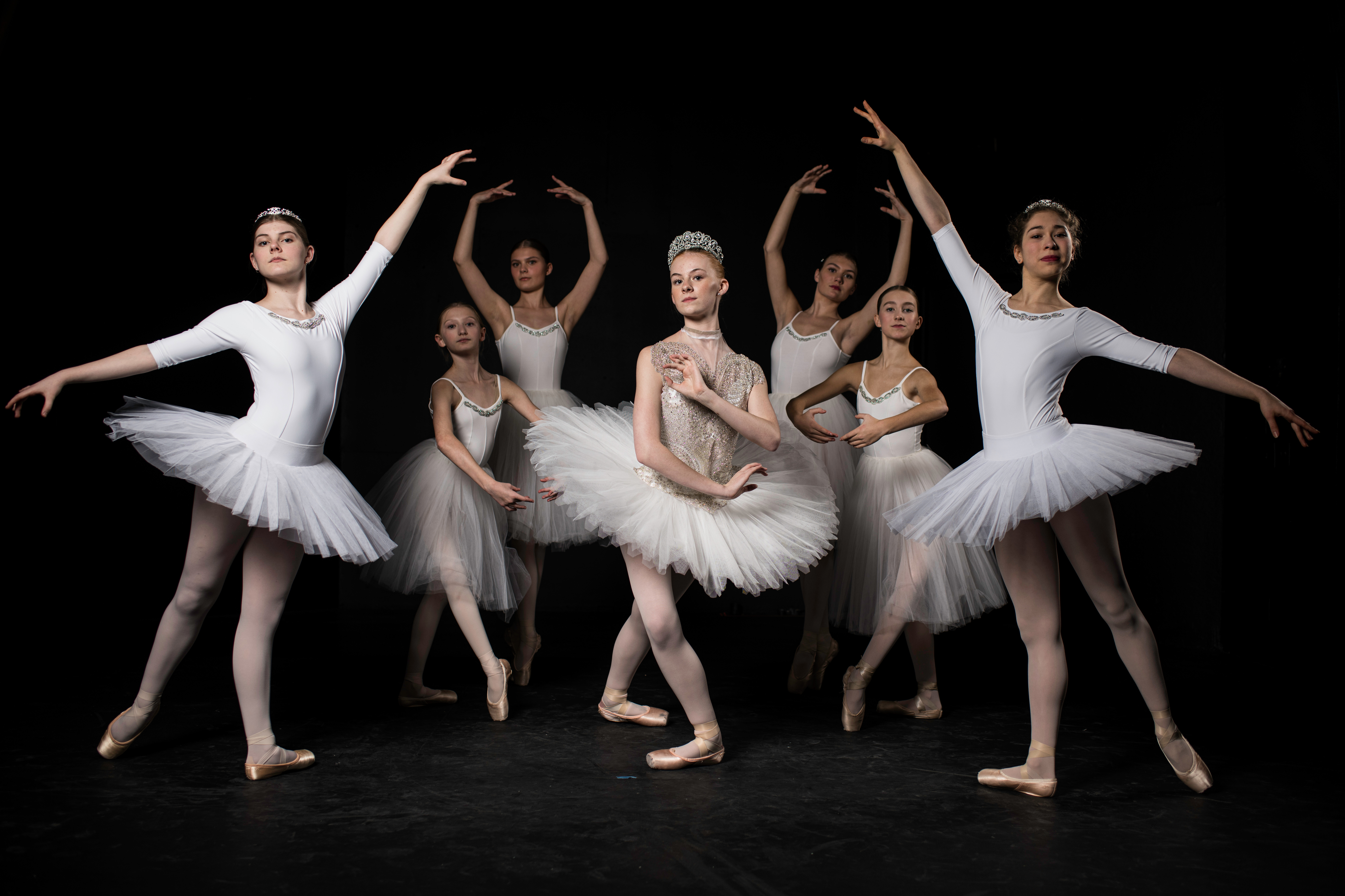 Youth dancers in white leotards and tutus pose against a black backdrop