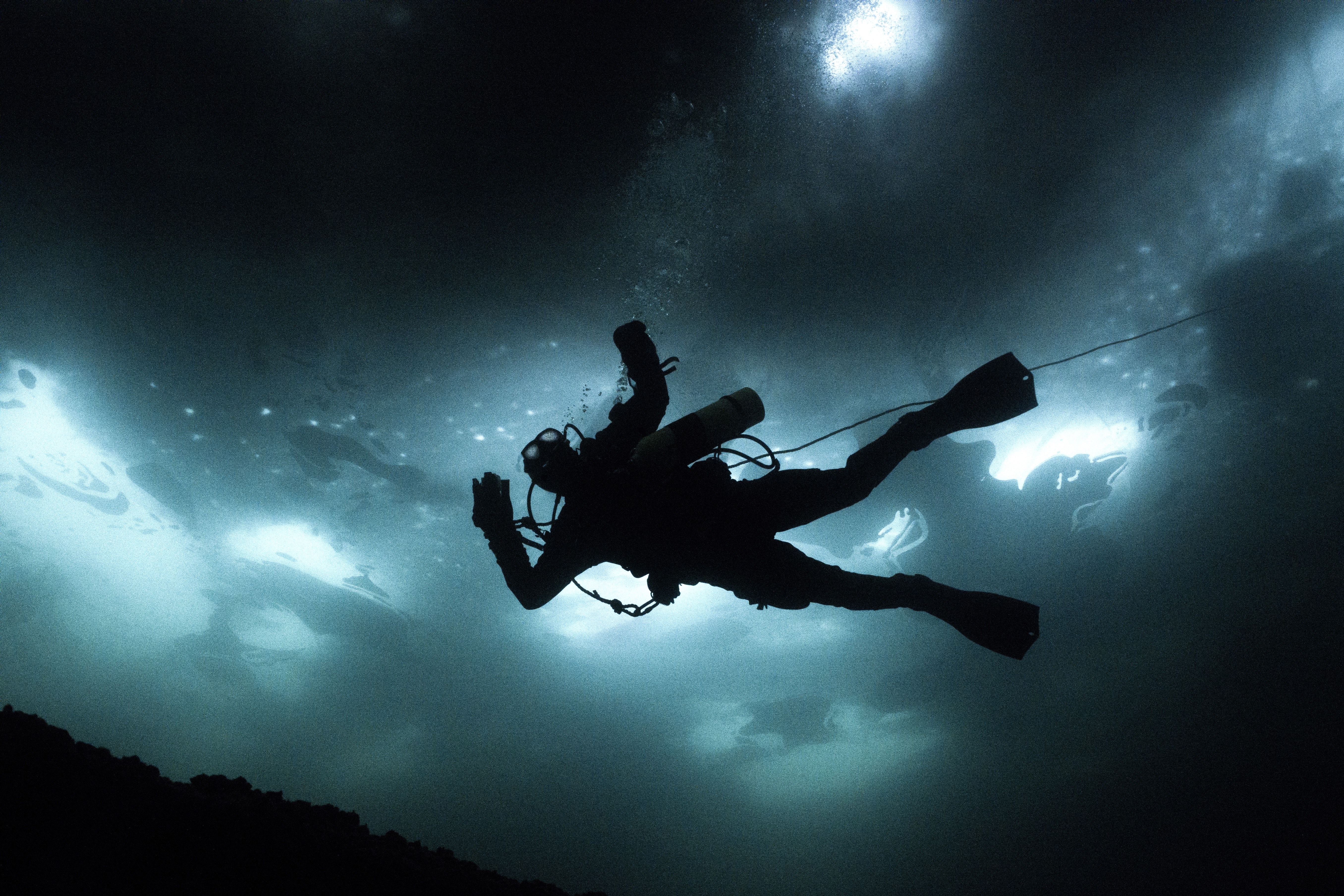 Underwater photograph of a scuba diver silhouetted against the light filtering through the surface above, with air bubbles rising and the diver’s gear clearly visible.