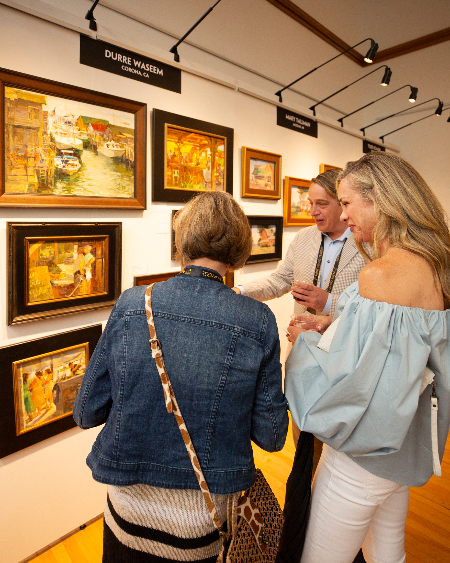 Two women and a man smile and point while they face a gallery wall lined with freshly painted plein air artworks.