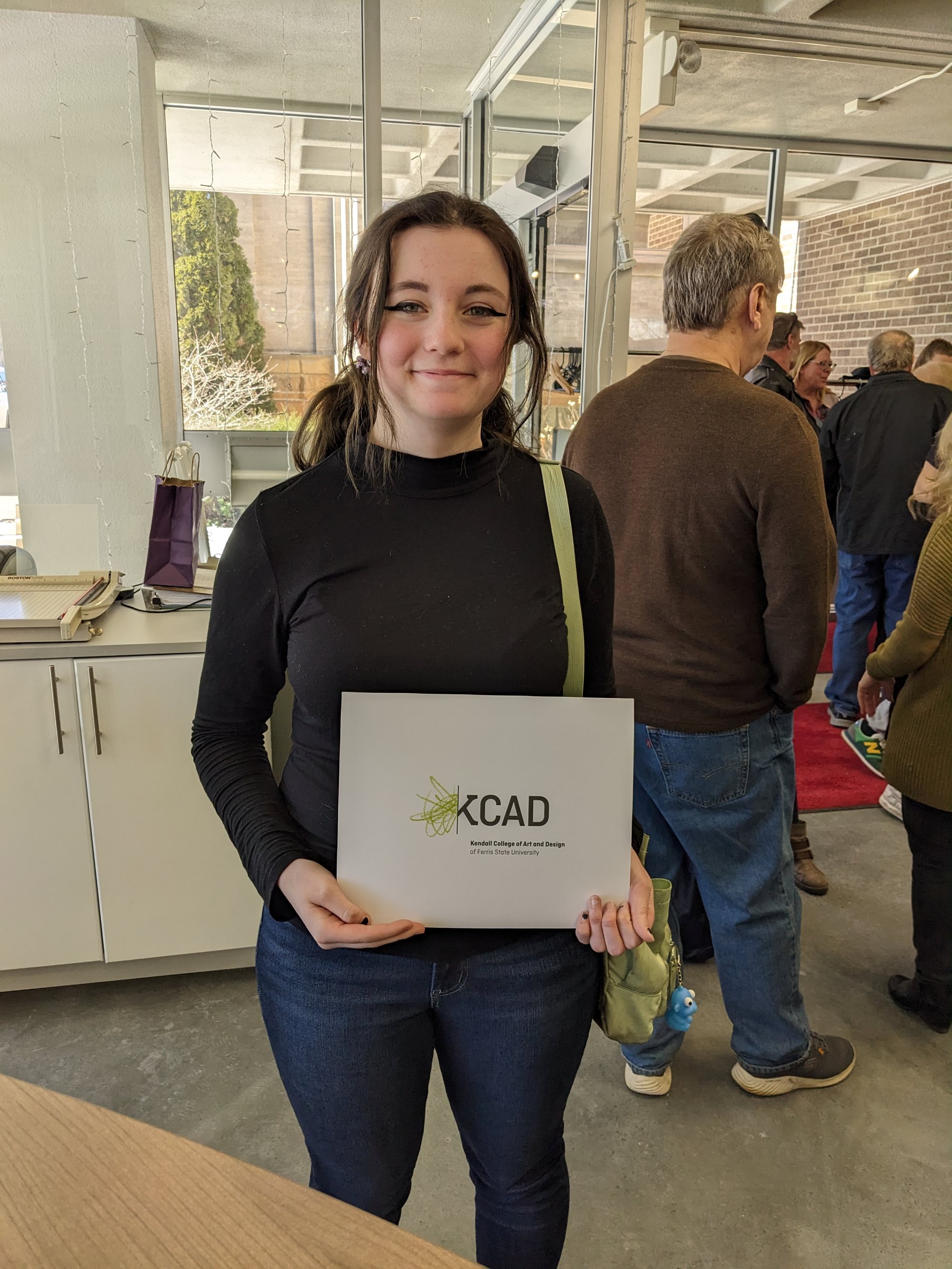 Brunette female student in brown sweater and jeans smiling while holding a white envelope with KCAD on the cover
